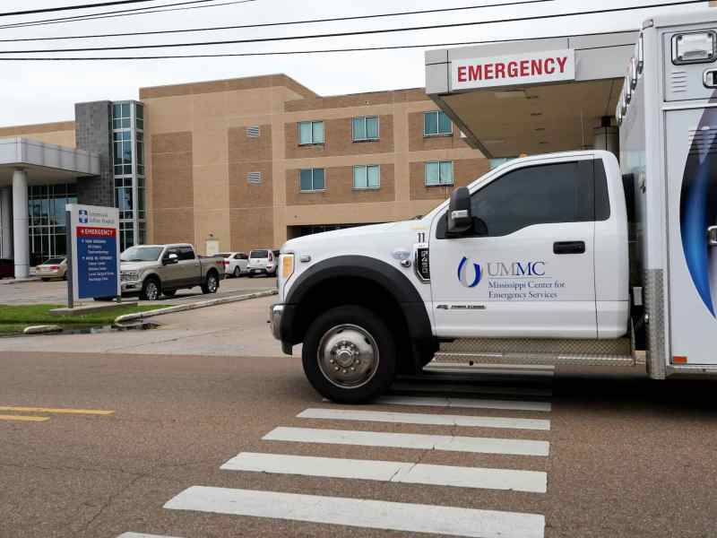 A light brown brick building labeled Greenwood Leflore Hospital, with a UMMC Emergency Services vehicle driving past