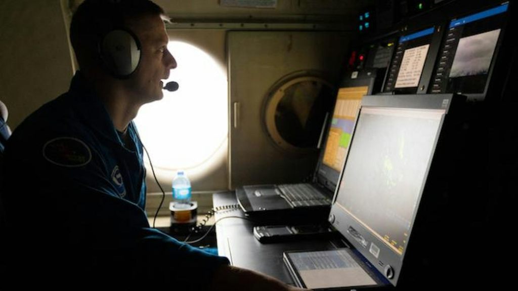 A scientists in a flight suit sits at a computer in an airplane talking on a headset